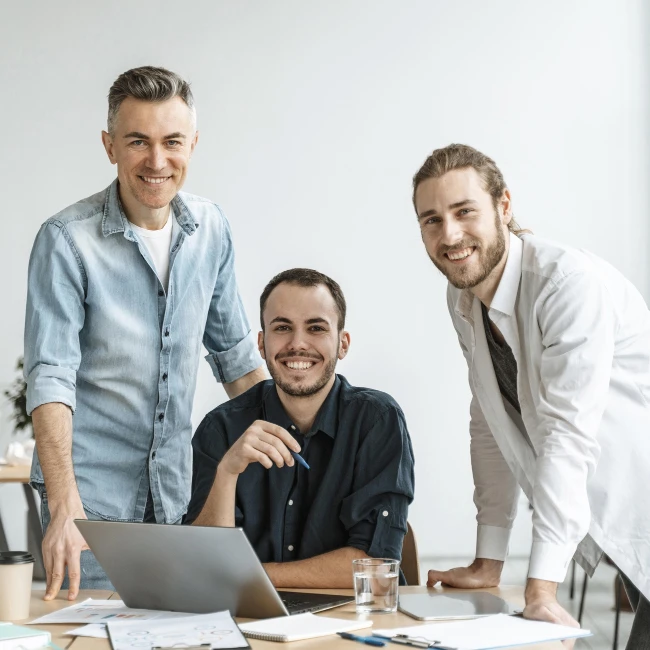 Trei bărbați zâmbind la birou, lucrând împreună la un laptop